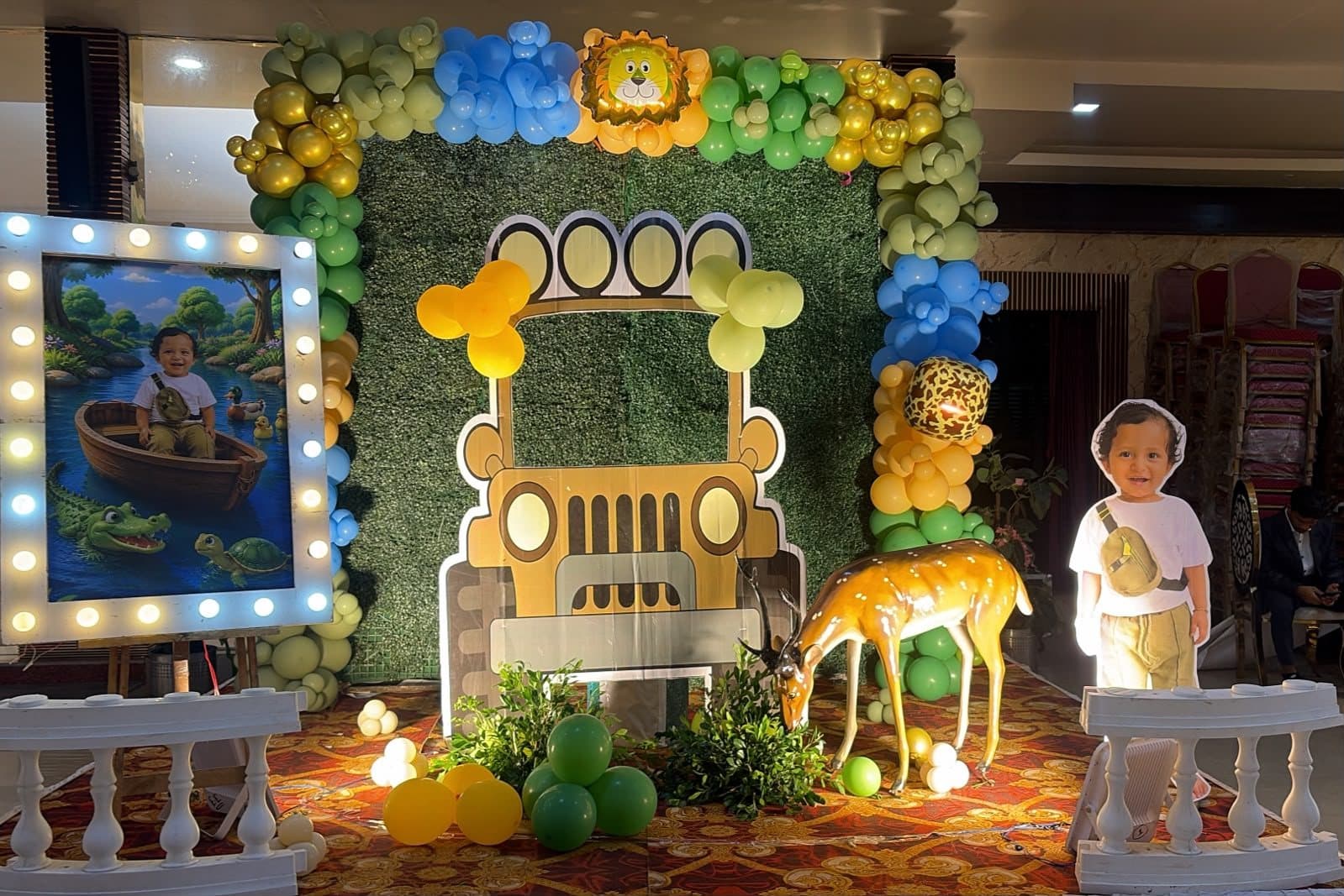 Indoor safari birthday backdrop with a jeep cutout on a green hedge wall, lion balloon and a life-size deer prop