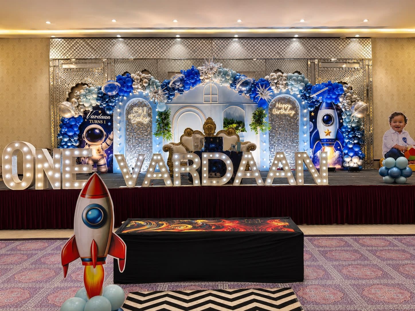 Space-themed first birthday stage with blue and silver balloon arch, rocket cutout and illuminated ONE VARDAAN marquee letters